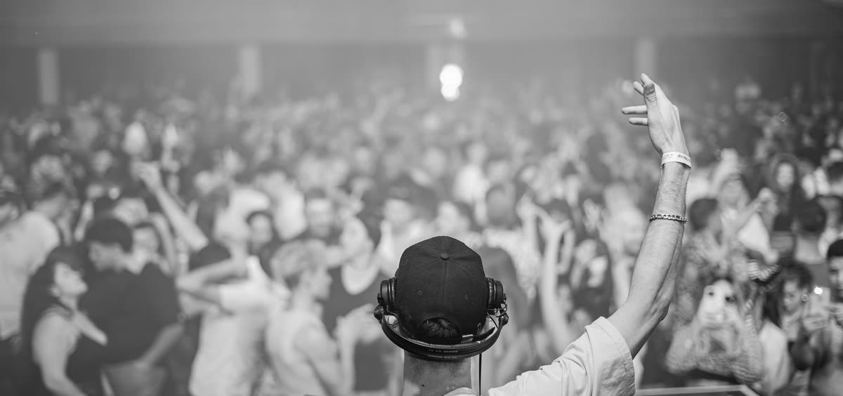 DJ seen from behind, arm raised to a packed nightclub crowd